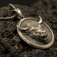 Load image into Gallery viewer, Bronze bull pendant necklace with an oxidized finish resting on textured volcanic rock, captured in a close-up premium studio composition.
