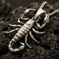 Load image into Gallery viewer, Bronze scorpion pendant necklace with an oxidized finish resting on textured volcanic rock, captured in a close-up premium studio composition.
