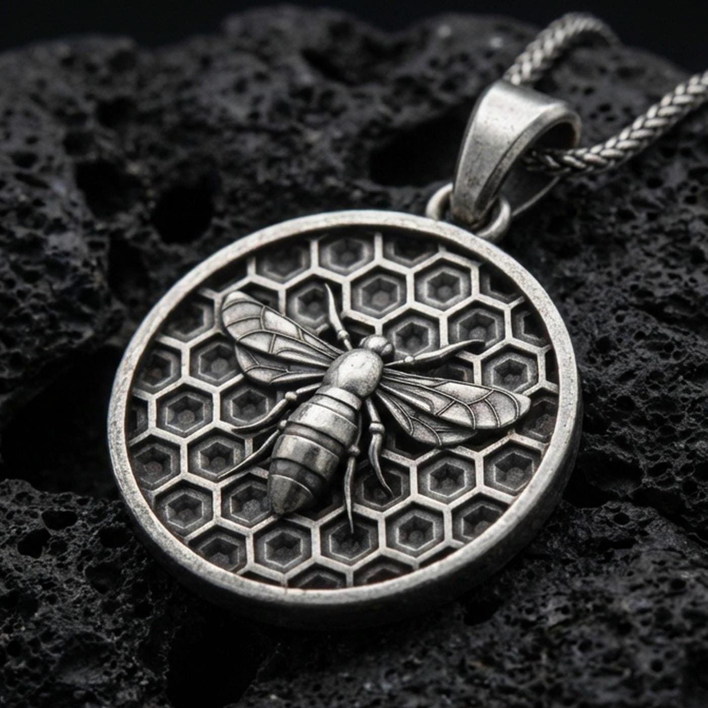 Sterling silver bee pendant necklace with an oxidized finish resting on textured volcanic rock, captured in a close-up premium studio composition.