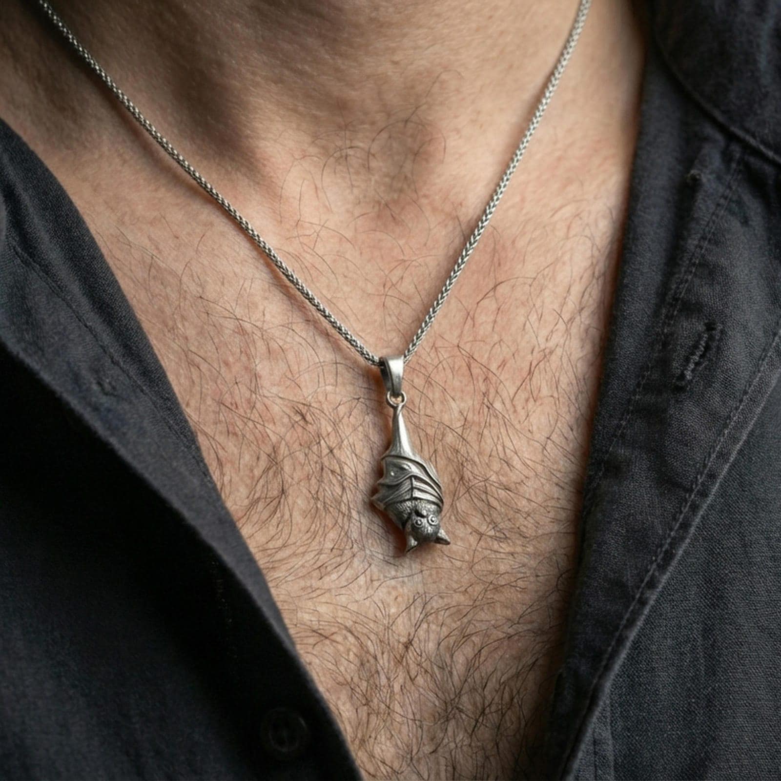 Oxidized hanging bat pendant necklace worn on the neck, styled in a dark lifestyle portrait with an open shirt and masculine presentation.