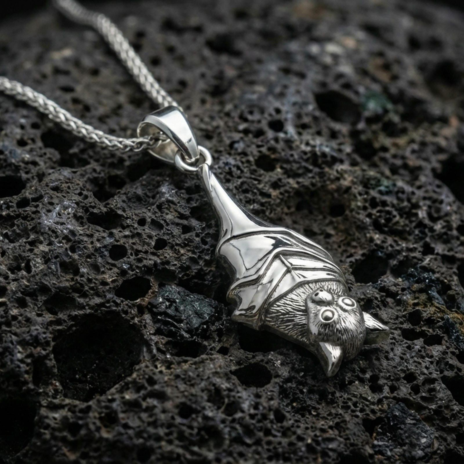 Polished silver hanging bat pendant necklace displayed on black volcanic rock, shown in a refined macro product shot with detailed wing wrap and sculpted bat face.