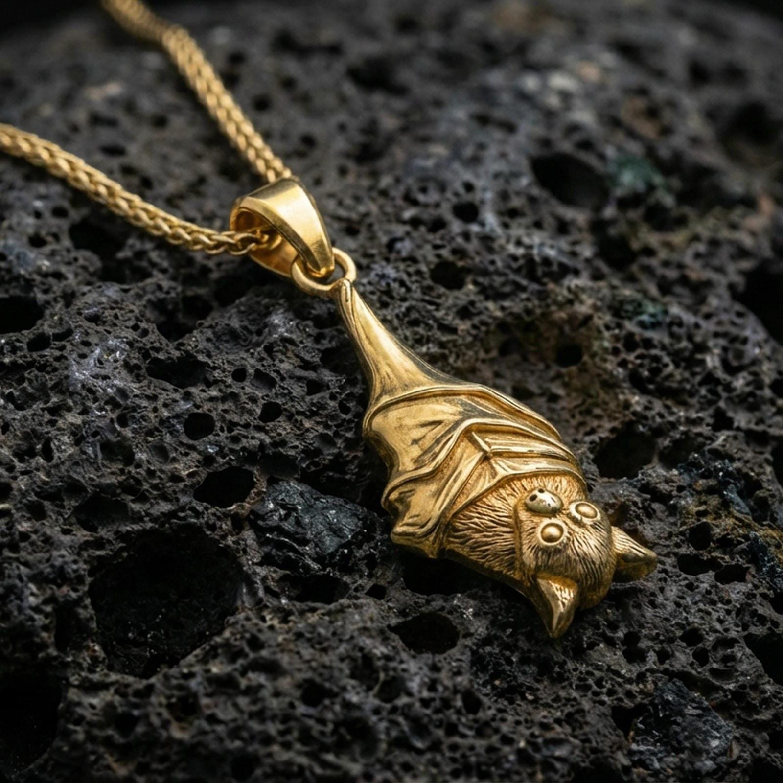 Gold plated hanging bat pendant necklace presented on porous volcanic rock, highlighting the gothic animal form in a luxurious close-up jewelry image.