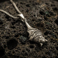 Load image into Gallery viewer, Bronze hanging bat pendant necklace with an oxidized finish resting on textured volcanic rock, captured in a close-up premium studio composition.
