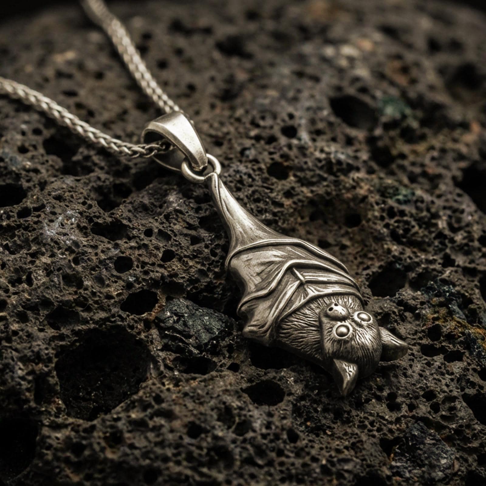 Bronze hanging bat pendant necklace with an oxidized finish resting on textured volcanic rock, captured in a close-up premium studio composition.