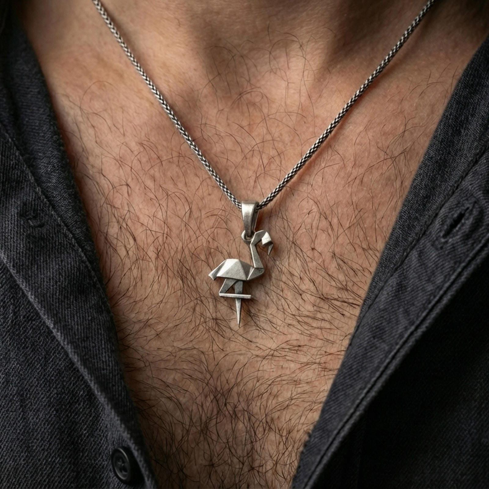 Oxidized origami flamingo pendant necklace worn on the neck, styled in a dark lifestyle portrait with an open shirt and masculine presentation.