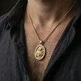 Load image into Gallery viewer, Gold plated Saint Francis necklace with oval saint portrait and cross motif worn on the neck in a premium lifestyle composition with classic Christian jewelry styling
