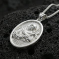 Load image into Gallery viewer, Polished silver Saint Francis medallion with detailed saint relief, halo, and cross motif placed on textured volcanic rock in a premium studio product shot
