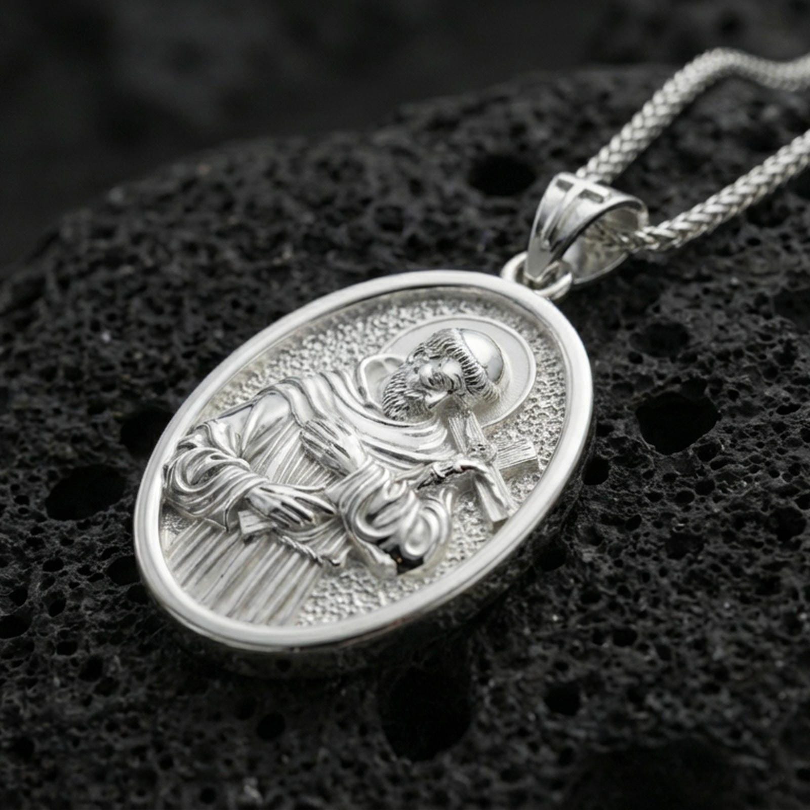 Polished silver Saint Francis medallion with detailed saint relief, halo, and cross motif placed on textured volcanic rock in a premium studio product shot