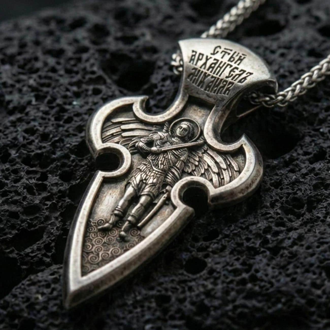 Oxidized Saint Michael cross pendant with winged archangel figure, engraved Cyrillic inscription, and elongated gothic silhouette displayed on dark volcanic rock in a close-up luxury jewelry composition