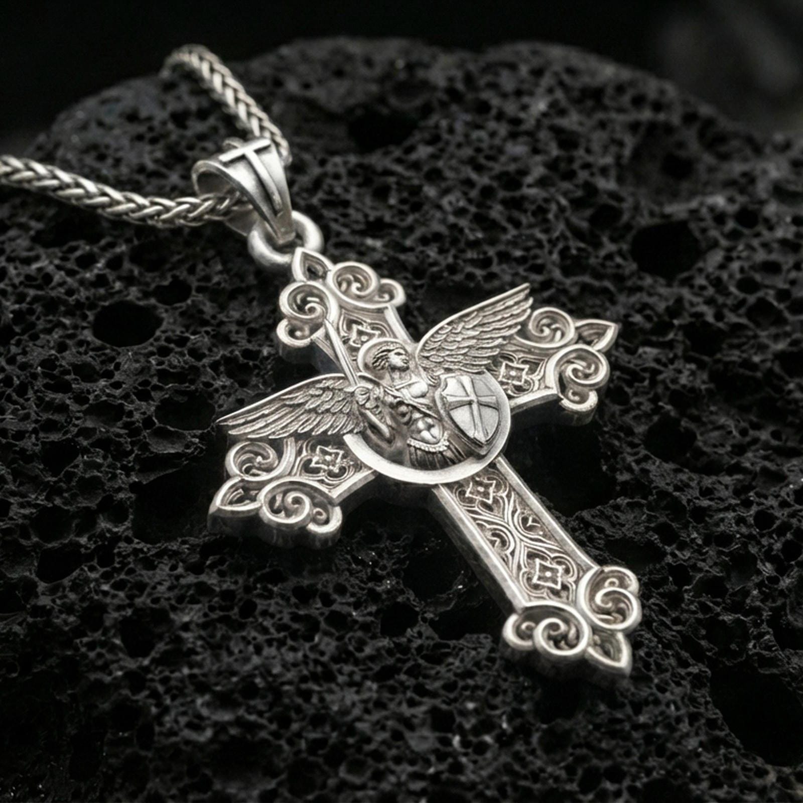 Polished silver Archangel Michael cross pendant with detailed angel warrior relief, decorative openwork flourishes, and shield motif placed on textured volcanic rock in a premium studio product shot