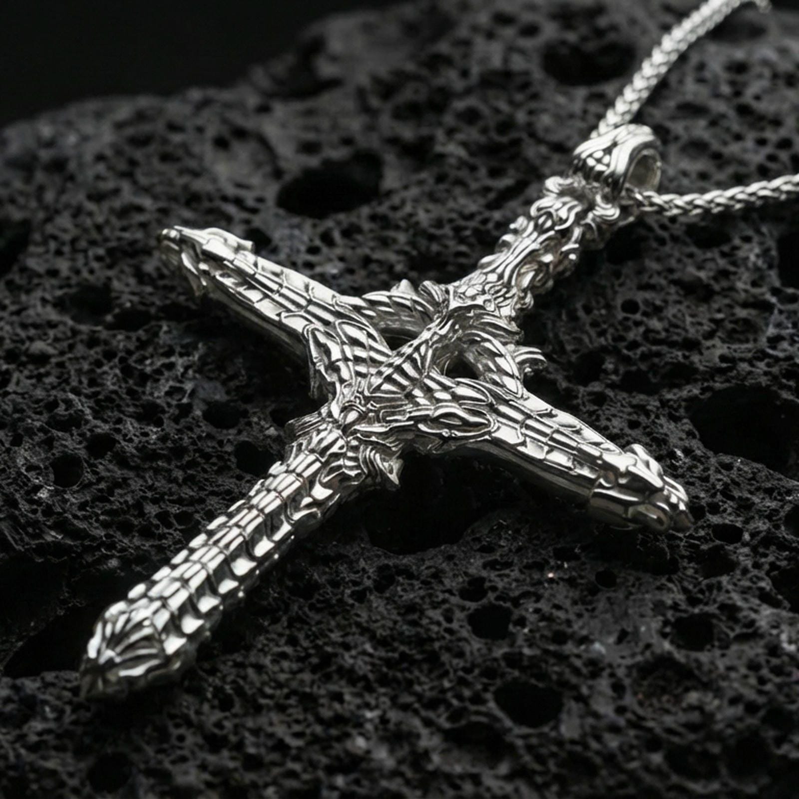 Polished silver gothic cross necklace with biomechanical relief, layered structural textures, and elongated cross silhouette placed on textured volcanic rock in a premium studio product shot