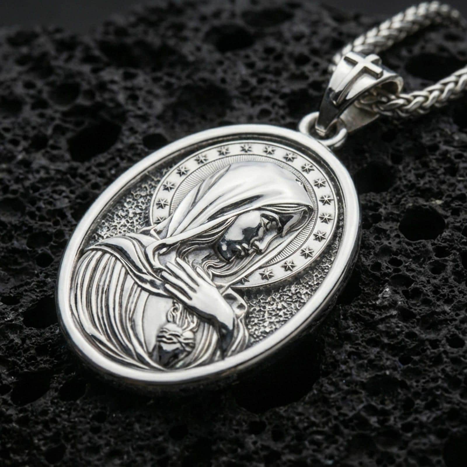 Polished silver Virgin Mary medal with veiled figure, star halo, and sacred heart detail placed on textured volcanic rock in a premium studio product shot