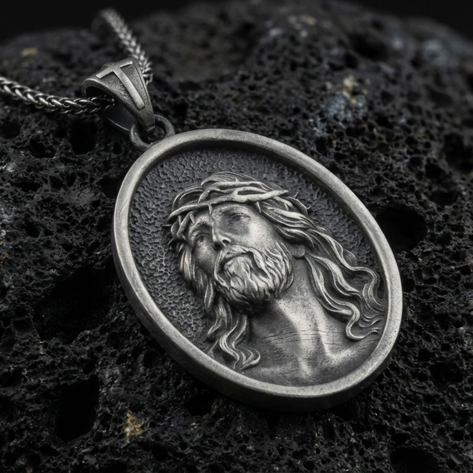 Oxidized Jesus Christ pendant with crown of thorns and detailed sacred portrait displayed on dark volcanic rock in a close-up luxury jewelry composition