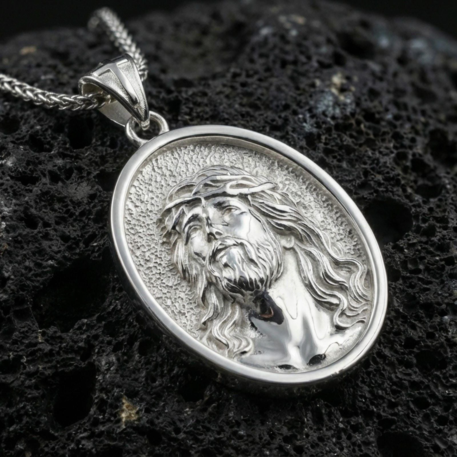 Polished silver Jesus medal with oval frame, crown of thorns, and sculpted devotional face placed on textured volcanic rock in a premium studio product shot
