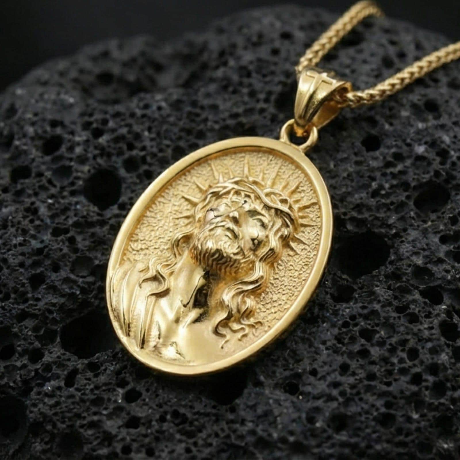 Gold plated Jesus Christ pendant with detailed crown of thorns portrait, radiant halo motif, and oval medallion design showcased on black volcanic rock in a refined macro jewelry image