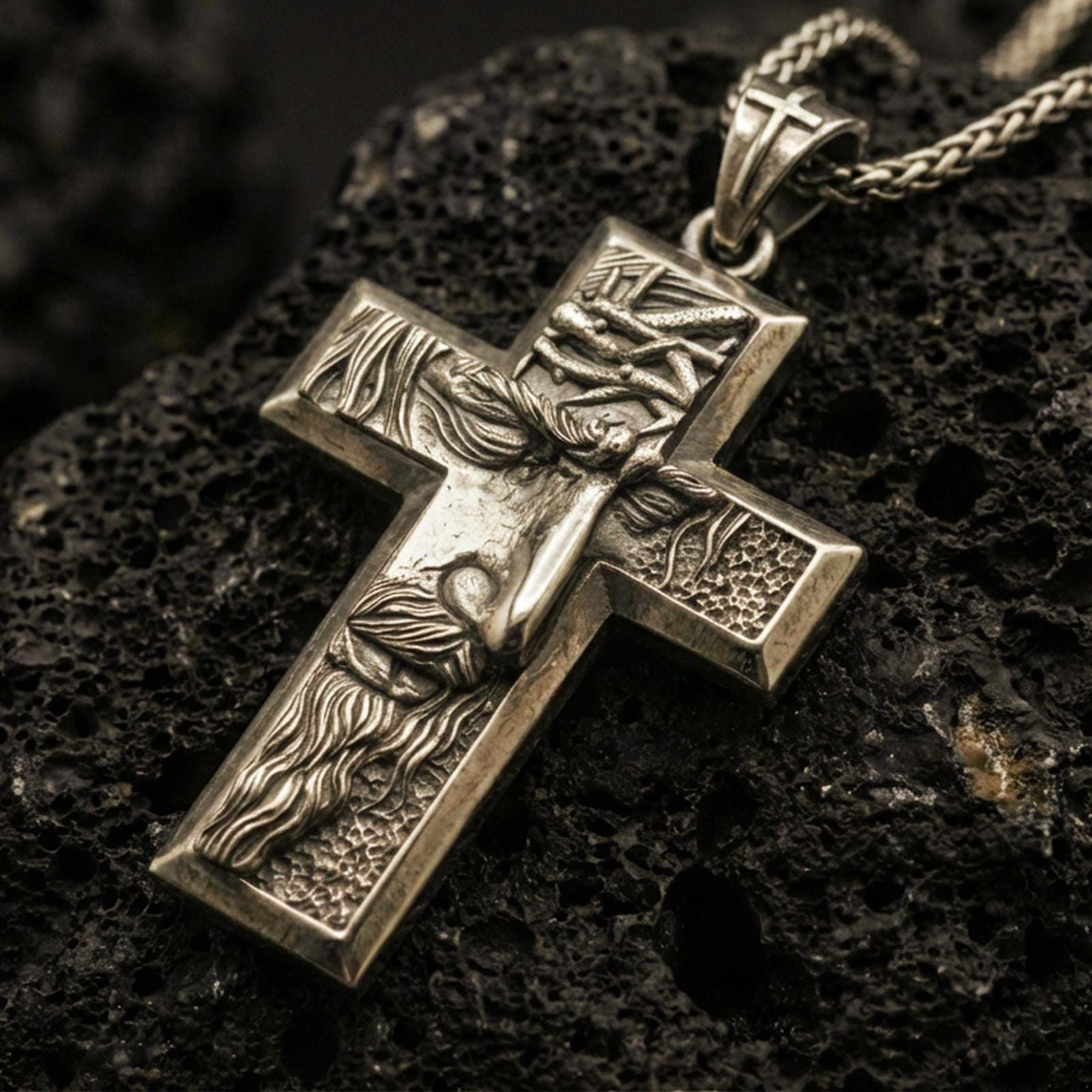 Bronze Jesus cross pendant with sculpted crucifix figure, crown of thorns, and textured relief displayed on dark volcanic rock in a close-up luxury jewelry composition