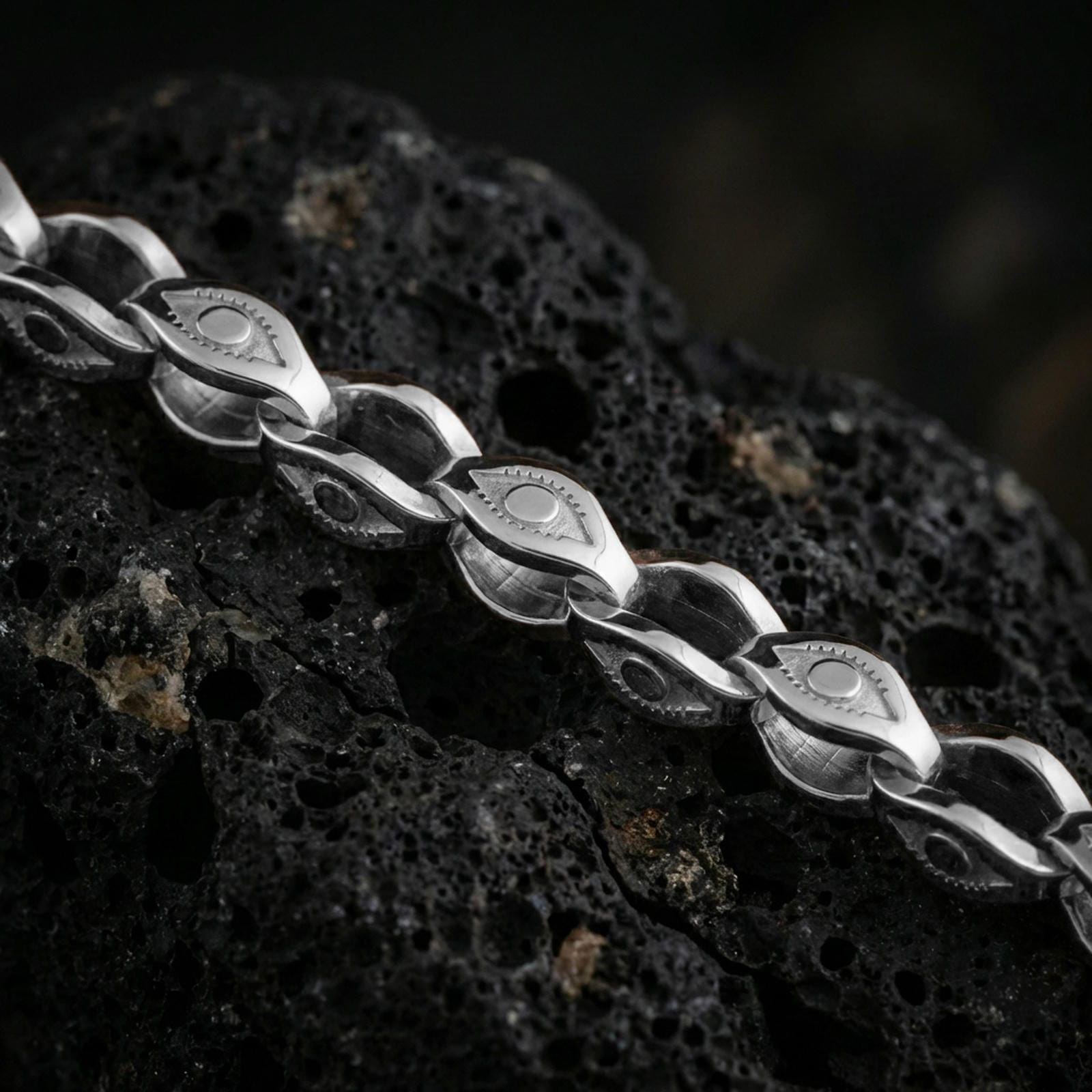 Polished silver evil eye link chain necklace displayed on black volcanic rock, shown in a refined macro product shot with engraved protective motif details.
