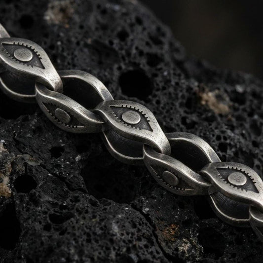 Sterling silver evil eye link chain necklace with an oxidized finish resting on textured volcanic rock, captured in a close-up premium studio composition.