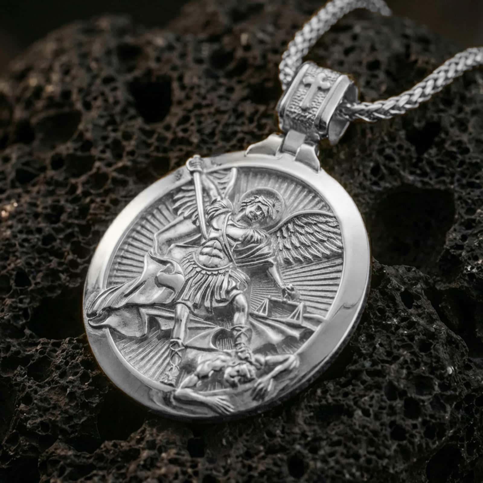Polished silver Saint Michael pendant necklace displayed on black volcanic rock, shown in a refined macro product shot with detailed archangel relief and symbolic battle scene.