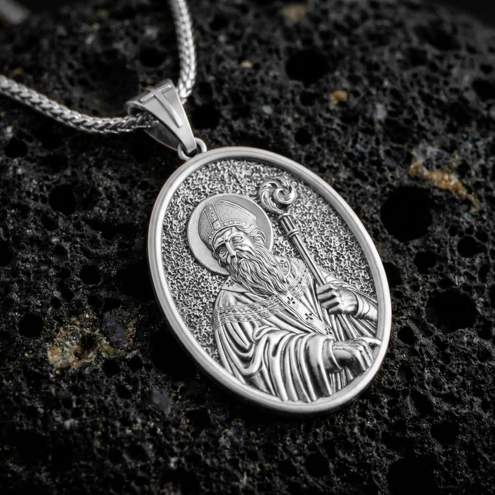 Polished silver Saint Patrick pendant necklace displayed on black volcanic rock, shown in a refined macro product shot with detailed bishop figure and pastoral staff relief.