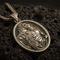 Load image into Gallery viewer, Bronze Saint Sebastian pendant necklace with an oxidized finish resting on textured volcanic rock, captured in a close-up premium studio composition.
