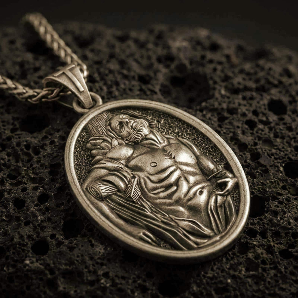 Bronze Saint Sebastian pendant necklace with an oxidized finish resting on textured volcanic rock, captured in a close-up premium studio composition.