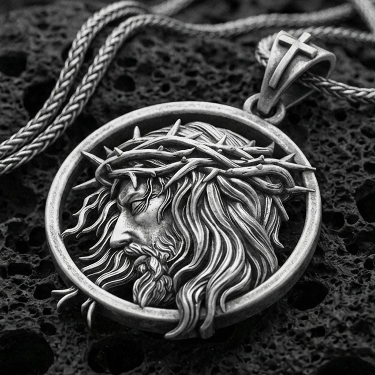 Oxidized Jesus crown of thorns pendant with side profile portrait and flowing hair detail displayed on dark volcanic rock in a close-up luxury jewelry composition