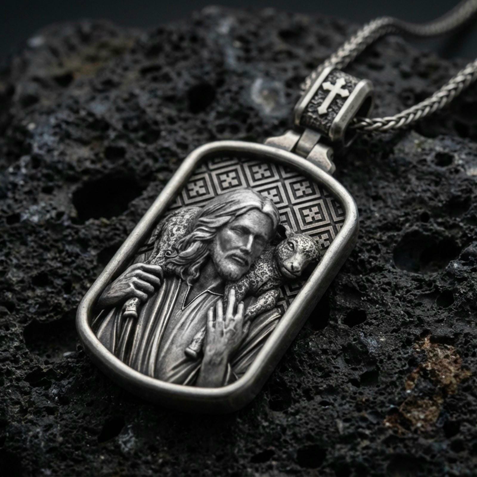 Oxidized Good Shepherd Jesus pendant with lamb over the shoulders and framed devotional portrait displayed on dark volcanic rock in a close-up luxury jewelry composition