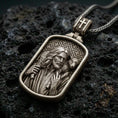 Load image into Gallery viewer, Bronze Good Shepherd Jesus pendant with lamb over the shoulders and framed devotional portrait displayed on dark volcanic rock in a close-up luxury jewelry composition
