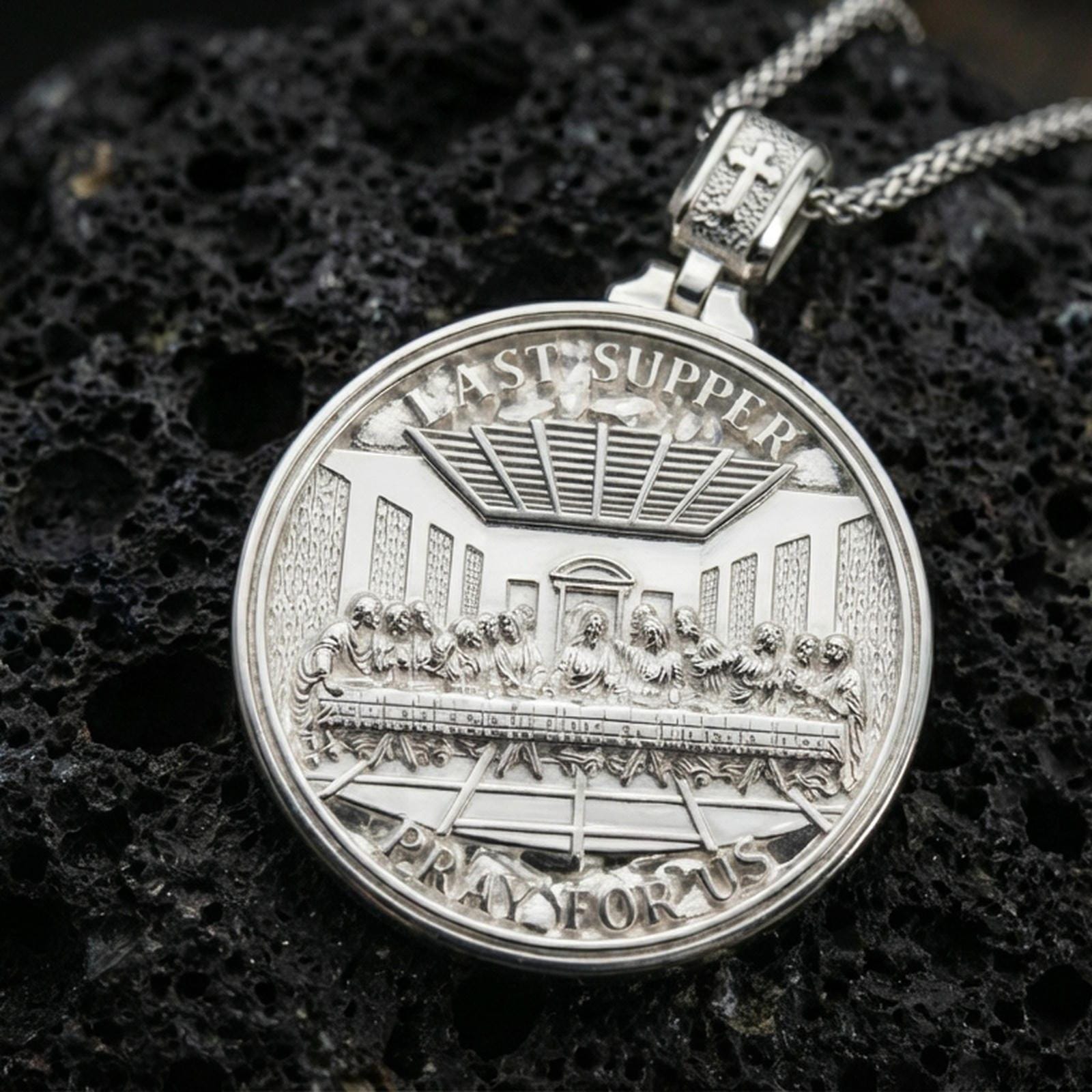 Polished silver Christian pendant with Last Supper relief on the front and Lord’s Prayer engraving on the back placed on textured volcanic rock in a premium studio product shot