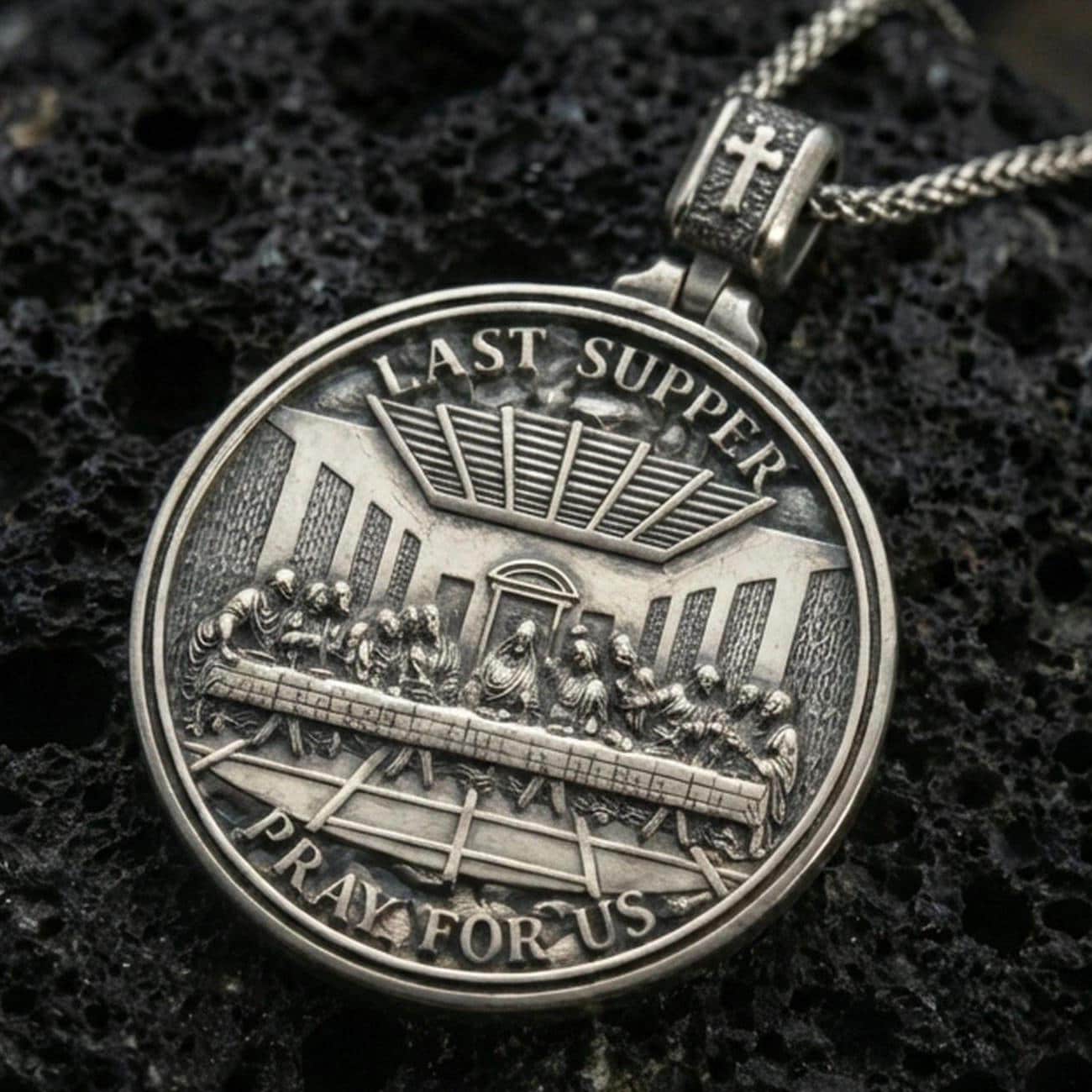 Oxidized Last Supper pendant with Pray for Us inscription, detailed round medallion design, and two-sided devotional concept displayed on dark volcanic rock in a close-up luxury jewelry composition