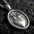 Load image into Gallery viewer, Oxidized Sacred Heart pendant with cross, flame detail, and oval devotional design displayed on dark volcanic rock in a close-up luxury jewelry composition
