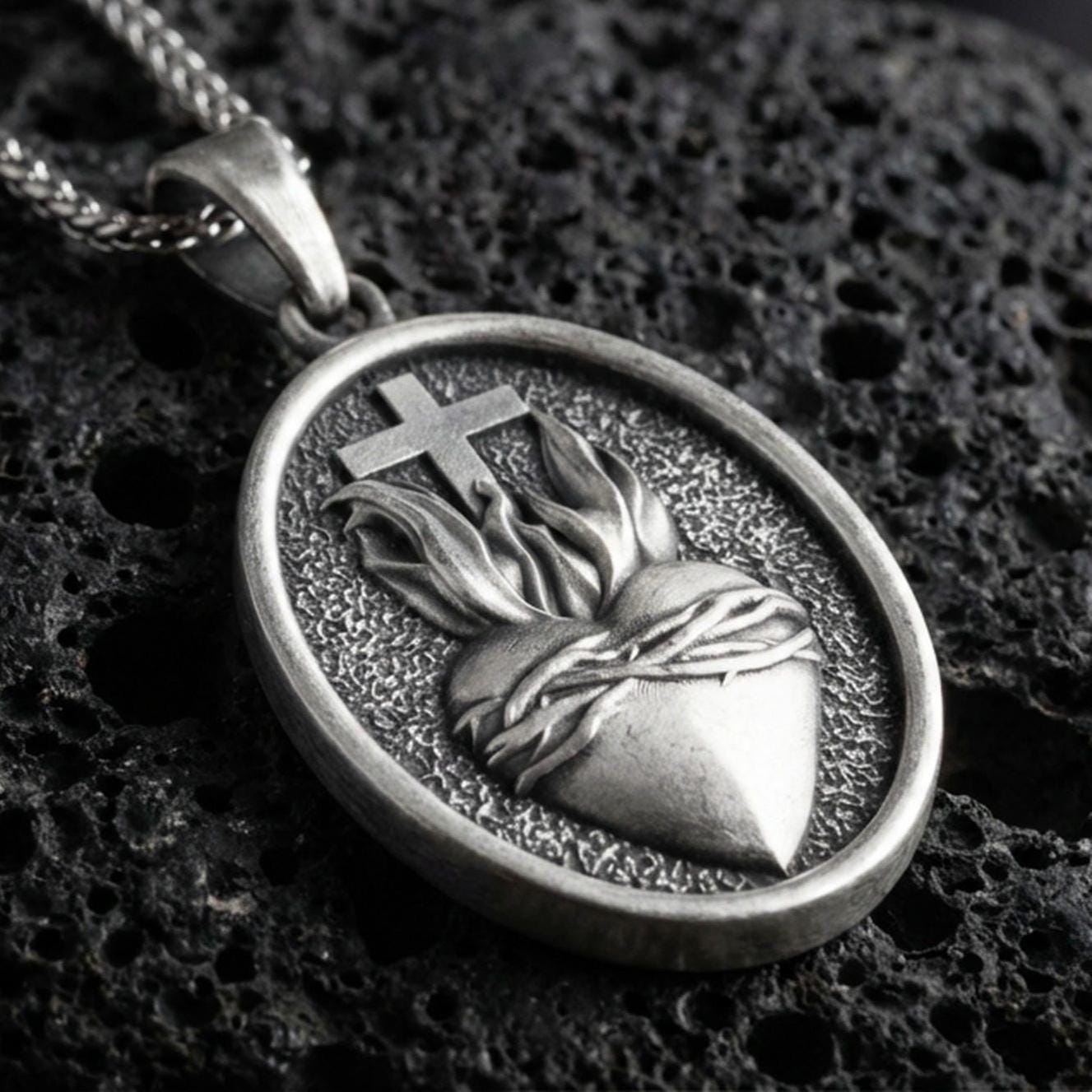 Oxidized Sacred Heart pendant with cross, flame detail, and oval devotional design displayed on dark volcanic rock in a close-up luxury jewelry composition
