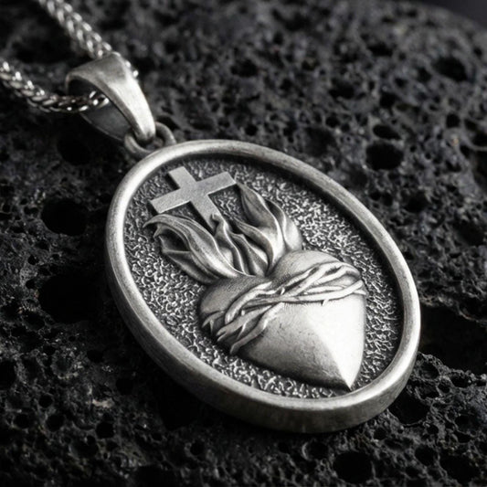 Oxidized Sacred Heart pendant with cross, flame detail, and oval devotional design displayed on dark volcanic rock in a close-up luxury jewelry composition