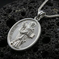 Load image into Gallery viewer, Polished silver Saint Dominic medallion with oval frame, robed saint relief, and engraved devotional lettering placed on textured volcanic rock in a premium studio product shot
