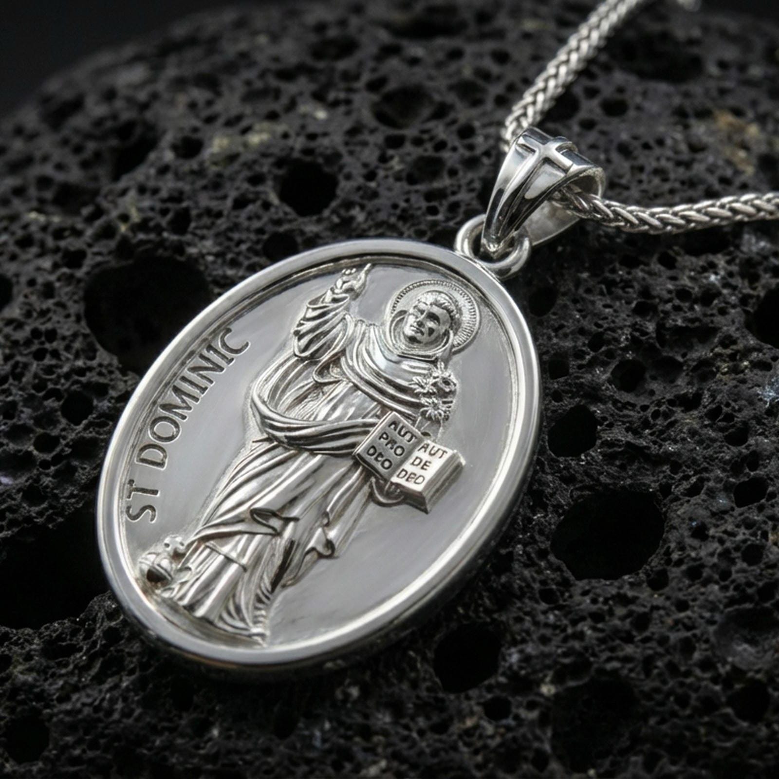 Polished silver Saint Dominic medallion with oval frame, robed saint relief, and engraved devotional lettering placed on textured volcanic rock in a premium studio product shot