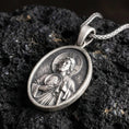 Load image into Gallery viewer, Oxidized Saint Jude pendant with haloed saint figure, medallion detail, and oval devotional portrait displayed on dark volcanic rock in a close-up luxury jewelry composition

