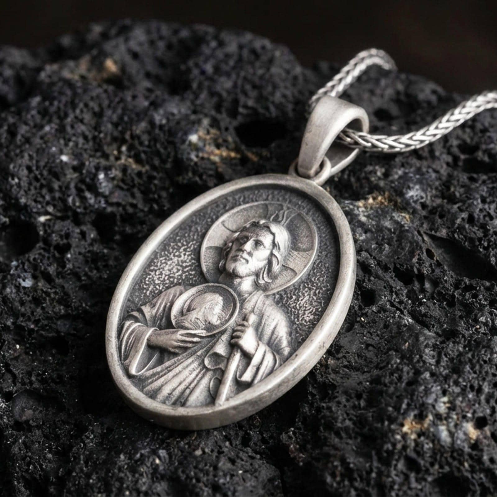 Oxidized Saint Jude pendant with haloed saint figure, medallion detail, and oval devotional portrait displayed on dark volcanic rock in a close-up luxury jewelry composition