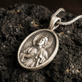 Load image into Gallery viewer, Bronze Saint Jude pendant with haloed saint figure, medallion detail, and oval devotional portrait displayed on dark volcanic rock in a close-up luxury jewelry composition
