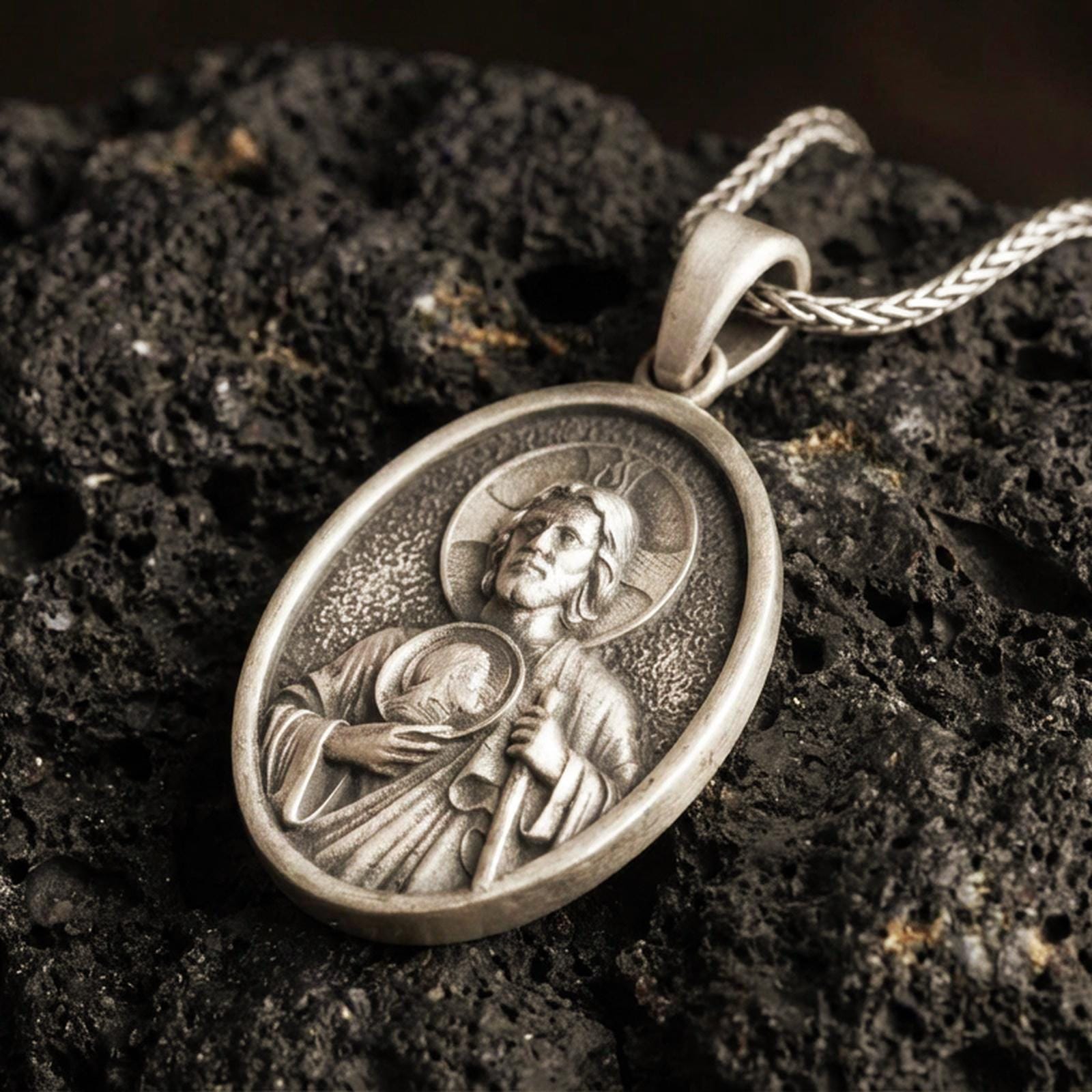 Bronze Saint Jude pendant with haloed saint figure, medallion detail, and oval devotional portrait displayed on dark volcanic rock in a close-up luxury jewelry composition