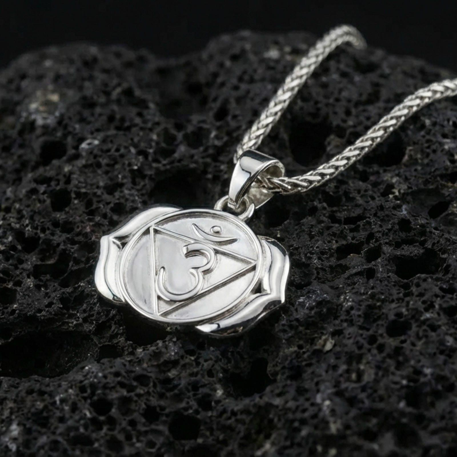 Polished silver chakra necklace with raised Om symbol, geometric triangle detail, and stylized lotus form placed on textured volcanic rock in a premium studio product shot