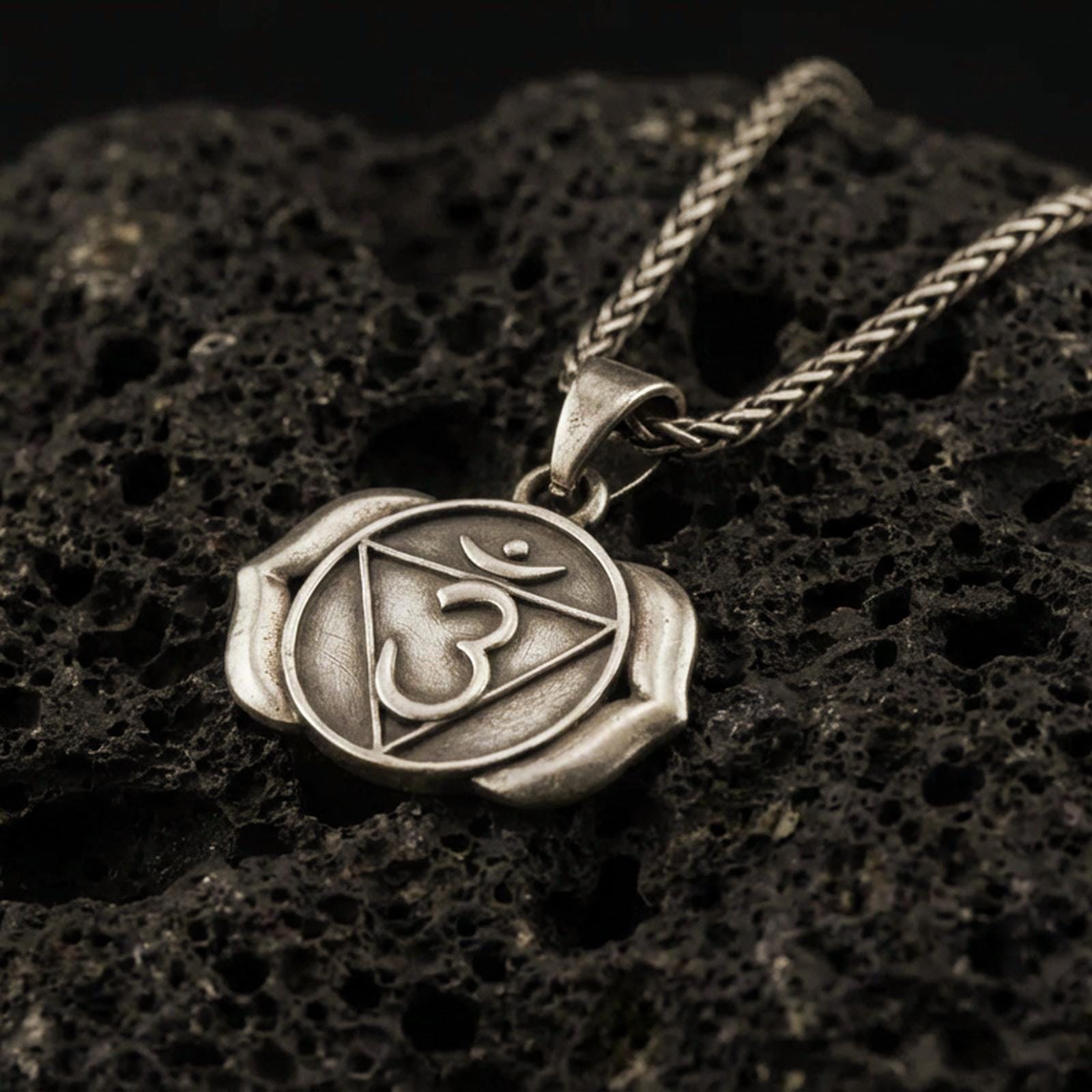 Bronze third eye chakra pendant with Om symbol, triangle motif, and two-petal lotus design displayed on dark volcanic rock in a close-up luxury jewelry composition