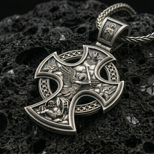 Oxidized Saint Michael cross pendant with winged archangel battle scene, openwork cross silhouette, and detailed heavenly relief displayed on dark volcanic rock in a close-up luxury jewelry composition