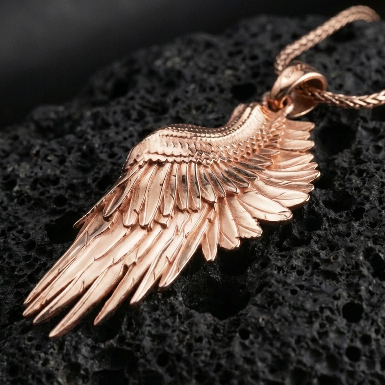Rose gold plated wing pendant with realistic feather relief, graceful elongated silhouette, and polished ornamental finish resting on volcanic rock in an elegant close-up product presentation