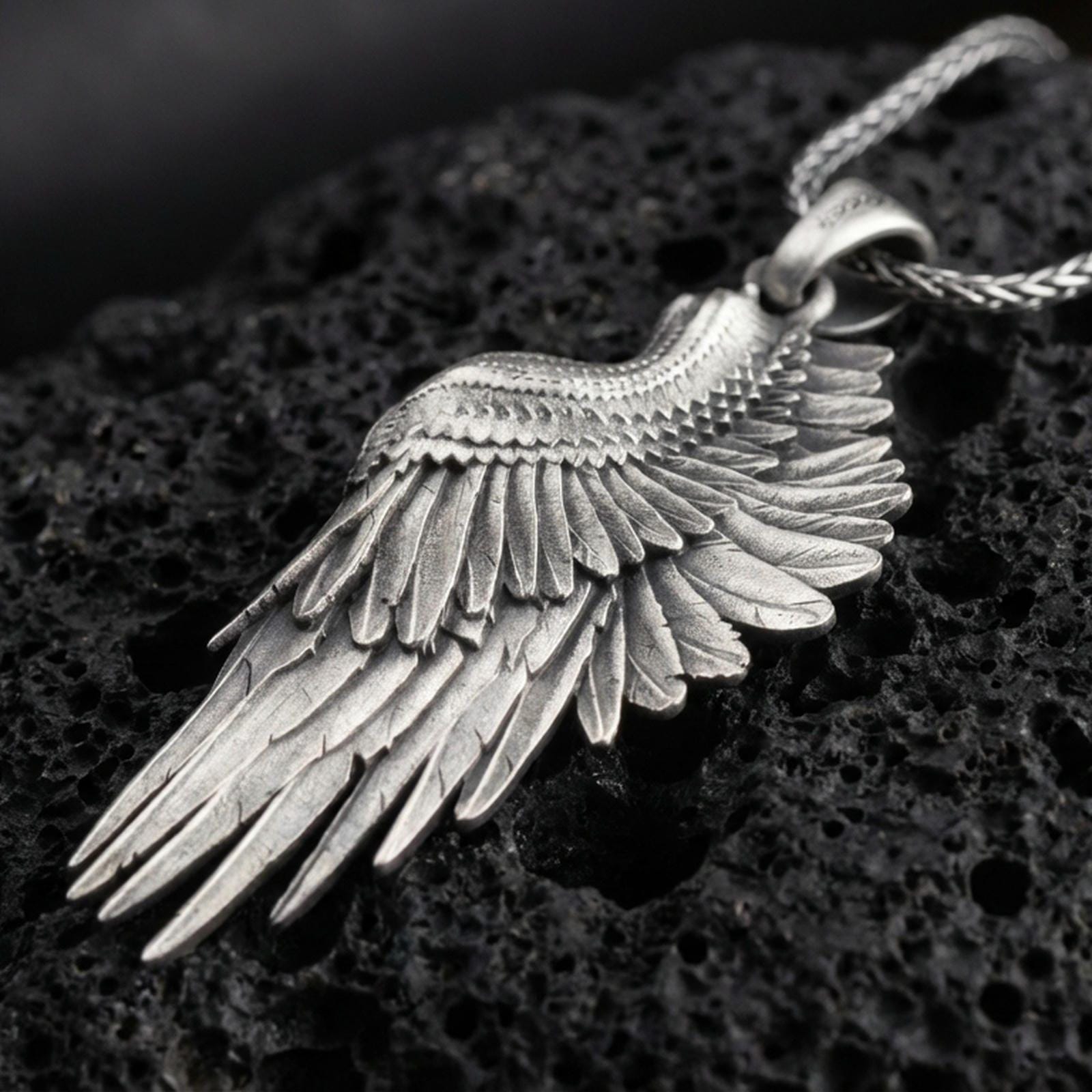 Oxidized angel wing pendant with layered feather detailing and elongated wing silhouette displayed on dark volcanic rock in a close-up luxury jewelry composition