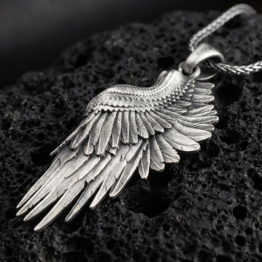 Oxidized angel wing pendant with layered feather detailing and elongated wing silhouette displayed on dark volcanic rock in a close-up luxury jewelry composition