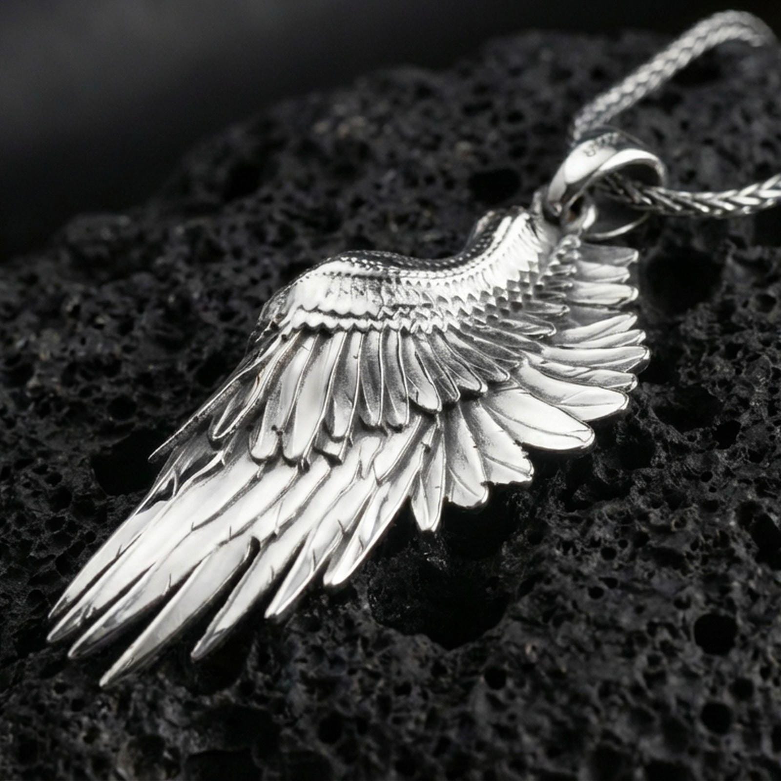 Polished silver wing necklace with sculpted feather texture, curved angel wing form, and refined dimensional relief placed on textured volcanic rock in a premium studio product shot