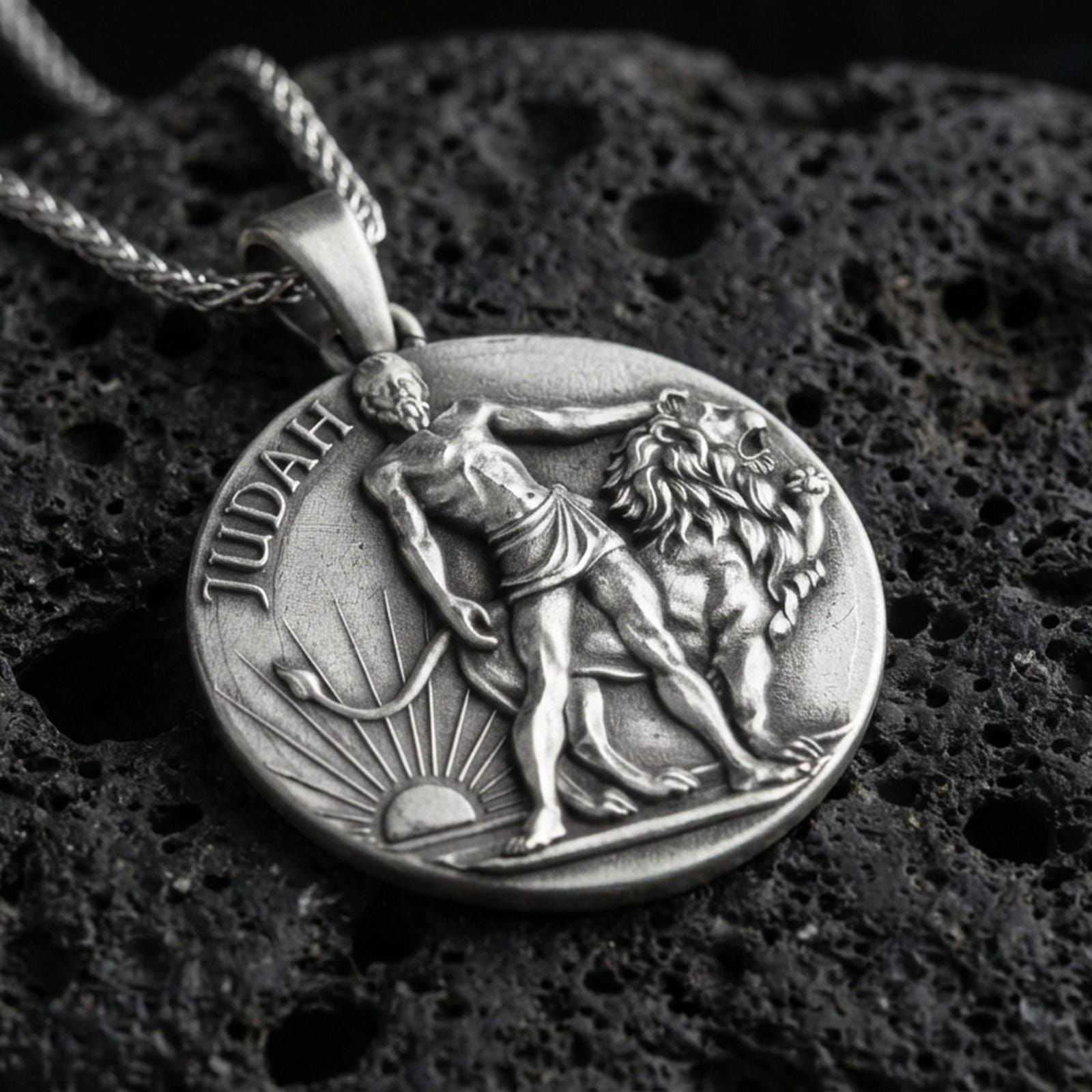 Oxidized Lion of Judah pendant with Judah inscription, lion figure, and sunrise motif displayed on dark volcanic rock in a close-up luxury jewelry composition