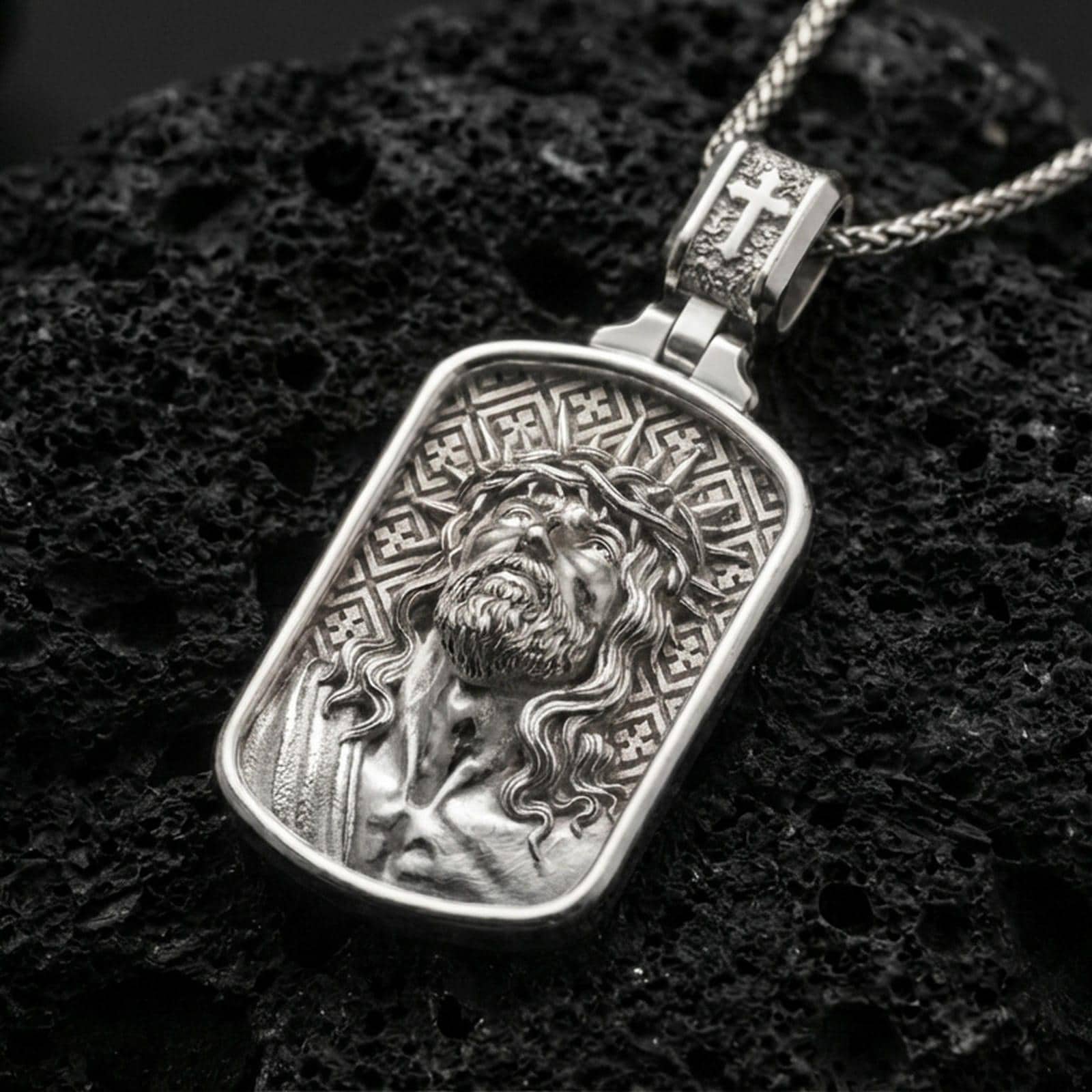 Polished silver Jesus medal with rounded rectangular frame, crown of thorns, and sculpted Christ portrait placed on textured volcanic rock in a premium studio product shot