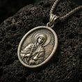 Load image into Gallery viewer, Bronze Saint Anastasia pendant with haloed saint figure, cross detail, and oval devotional portrait displayed on dark volcanic rock in a close-up luxury jewelry composition
